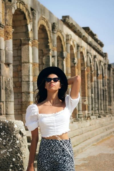 A tourist in a sunhat stands before the ancient Roman archways and pillars at the Volubilis historical site, showcasing the architectural wonder and comfortable exploration on the Fes day trip.