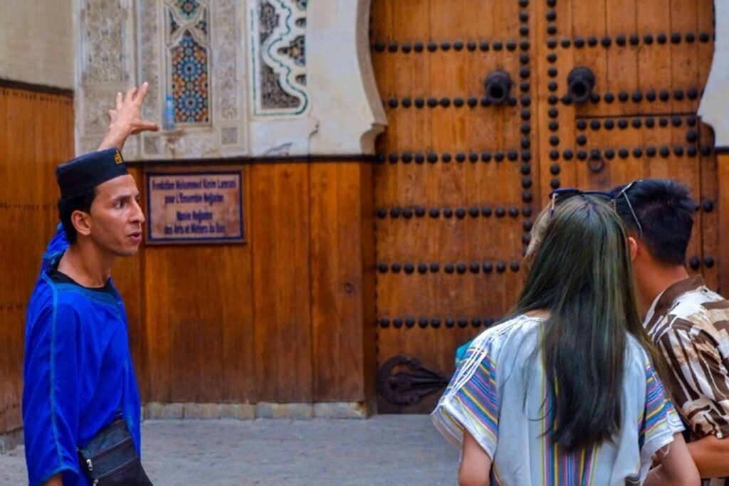 A professional tour guide wearing traditional attire explains landmarks to guests in front of an ornate wooden door in Fes, reflecting a personalized and outstanding guided city tour.