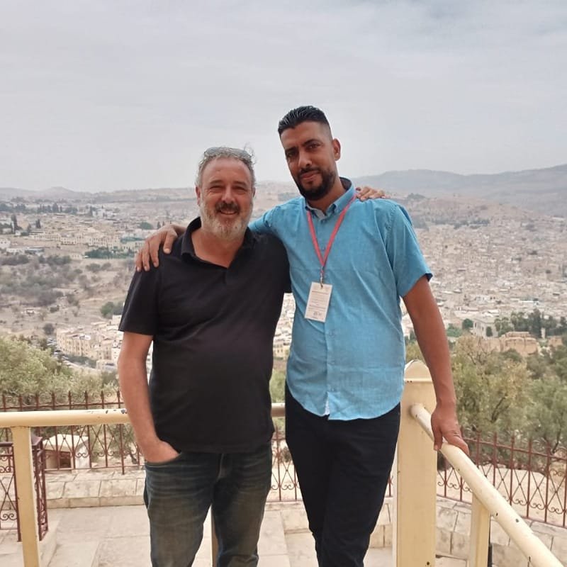 A friendly, professional local Fes guide with a badge standing next to a happy male tourist, posing against a panoramic view of the Fes Medina.