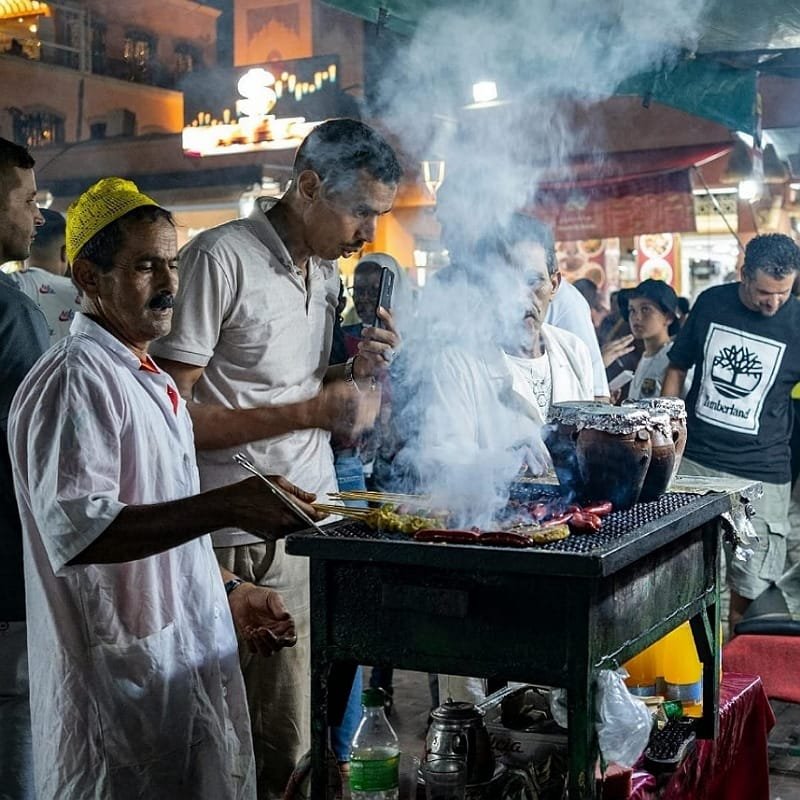 A Moroccan street food vendor grilling fresh skewers and kebabs on a smoky grill at night, surrounded by locals and tourists, highlighting a vibrant culinary tour experience.