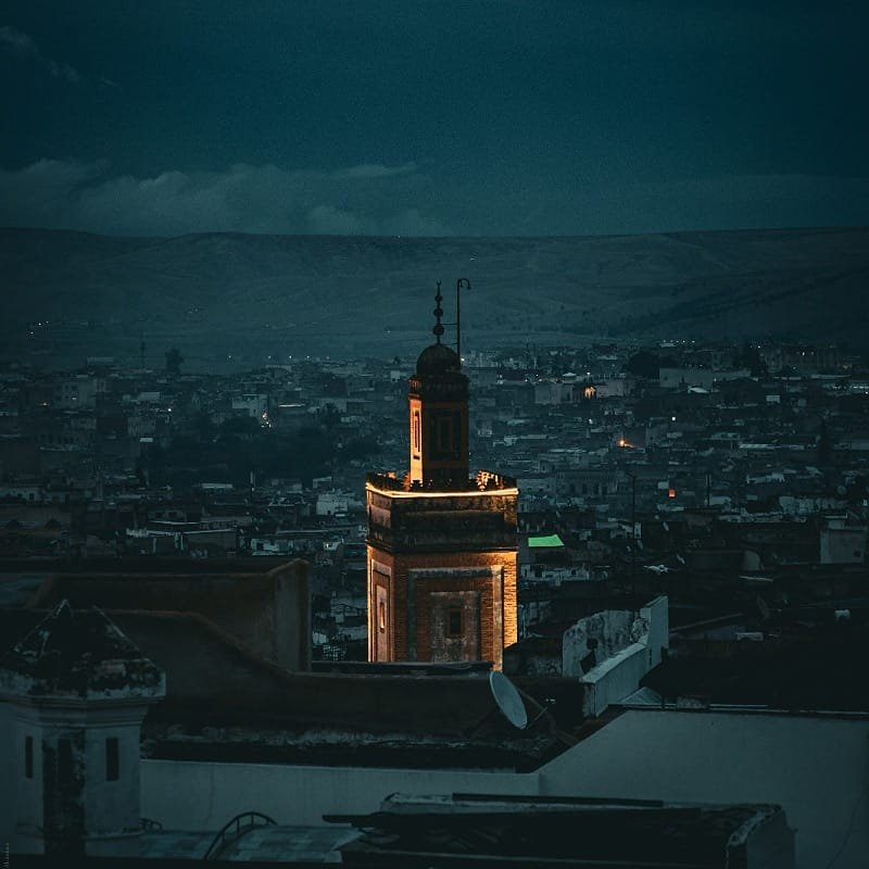 A beautifully illuminated minaret towering over the dark, sprawling Fes Medina skyline at night, with the silhouette of the surrounding hills under a dark sky, capturing the mystical atmosphere of a night tour.