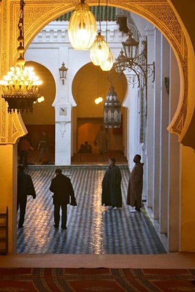 A warm, dimly lit view through a traditional Moroccan archway showing hanging lanterns and Zellige tiles in a historical building courtyard at night. Optimized for Fes cultural night tours.