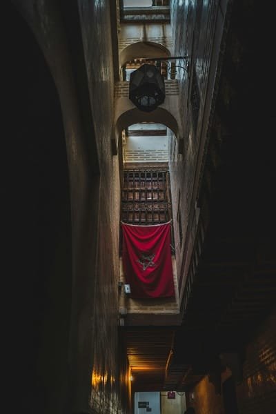 A vertical view of a narrow, old alleyway in the medina of Fez, dimly lit with a Moroccan flag hanging. It captures the authenticity and mystique of Fez's nighttime strolls.