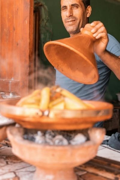A local Fes vendor lifting the conical lid of a traditional clay Tagine dish, showing the hot steam and fresh ingredients being cooked over charcoal, a central piece of the Fes Food Tour.