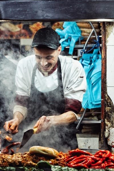 A happy Fes street food vendor in a chef's uniform cooking sausages and local food on a grill, highlighting the fresh, authentic, and passionate side of the Fes culinary tour.
