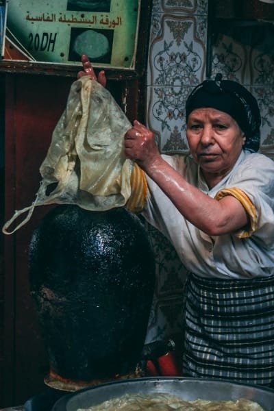 A local Fes woman preparing thin Pastilla or Msemen dough sheets using a traditional hot pot, showcasing the authentic, artisanal preparation methods of Fes street food.