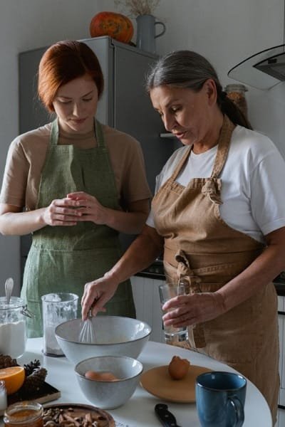Two women, one older (expert) and one younger (student), preparing batter and ingredients with aprons in a kitchen, representing the hands-on and intergenerational learning in a Fes cooking class.