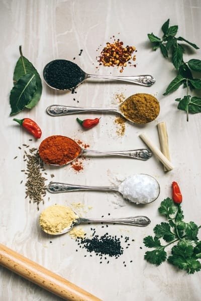 A vertical flat lay image showing various colorful Moroccan spices (paprika, cumin, turmeric, chili flakes) and fresh herbs essential for Tagine and Harira recipes in the Fes cooking class.