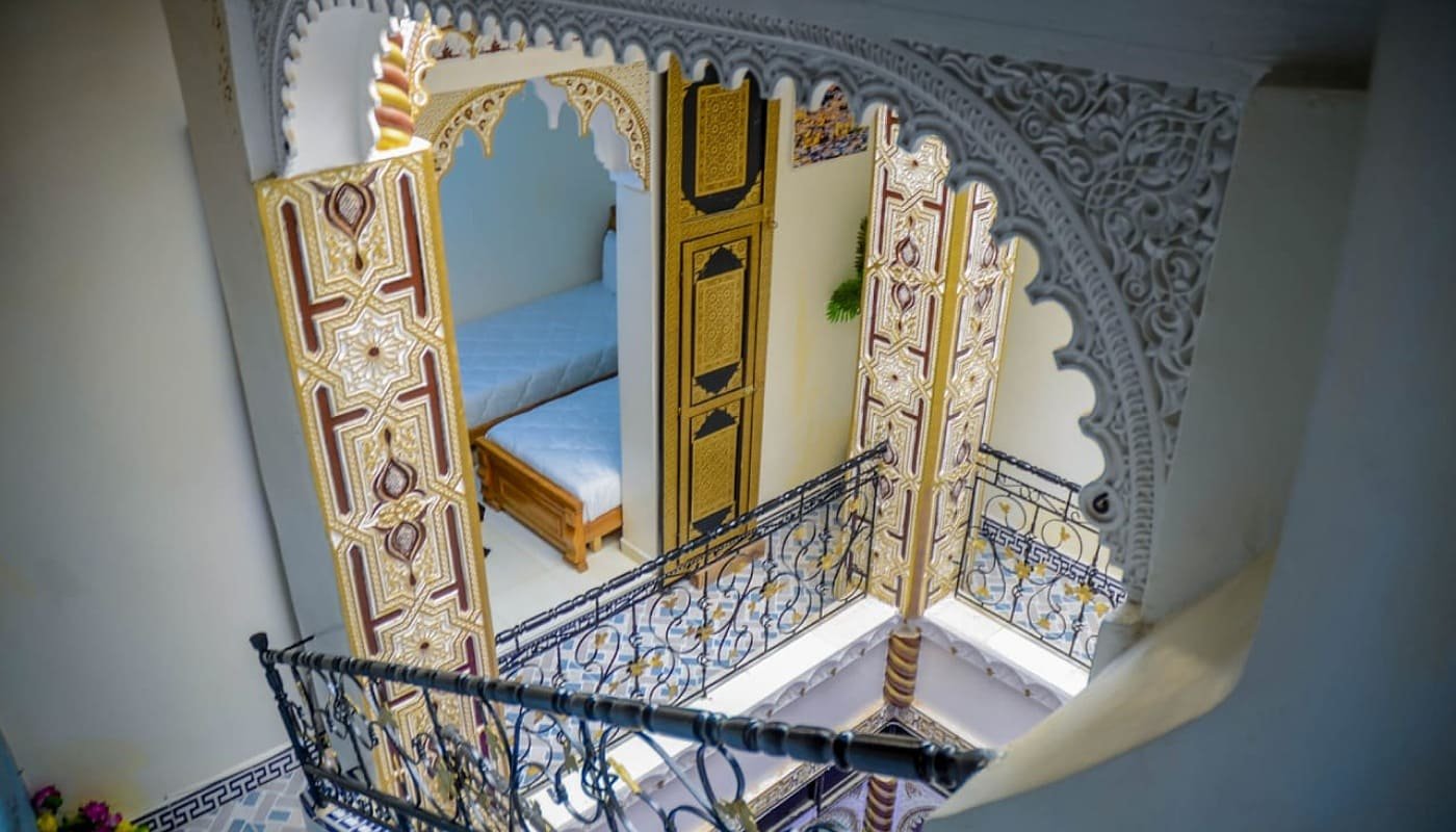 High-angle interior shot of LA CASA DAR RAYAN guesthouse, showcasing the intricate Moroccan archways, detailed gold and black banisters, and a glimpse into a traditional bedroom. Perfect for Fes luxury riad.
