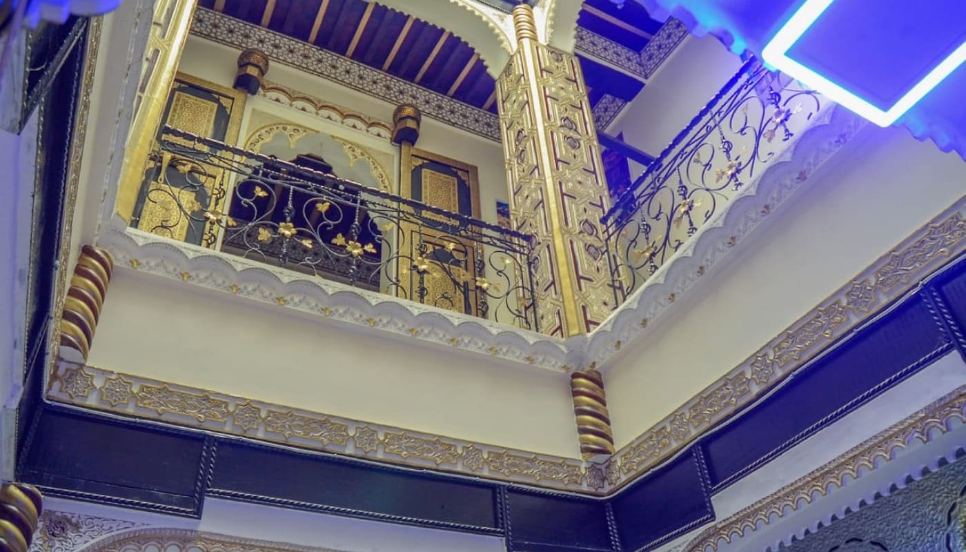 A high-angle photo of LA CASA DAR RAYAN's interior courtyard, showing ornate gold and black Moroccan carvings (gabs/stucco), traditional wooden ceilings, and wrought-iron balconies. Highlights the luxury Riad experience in Fes.