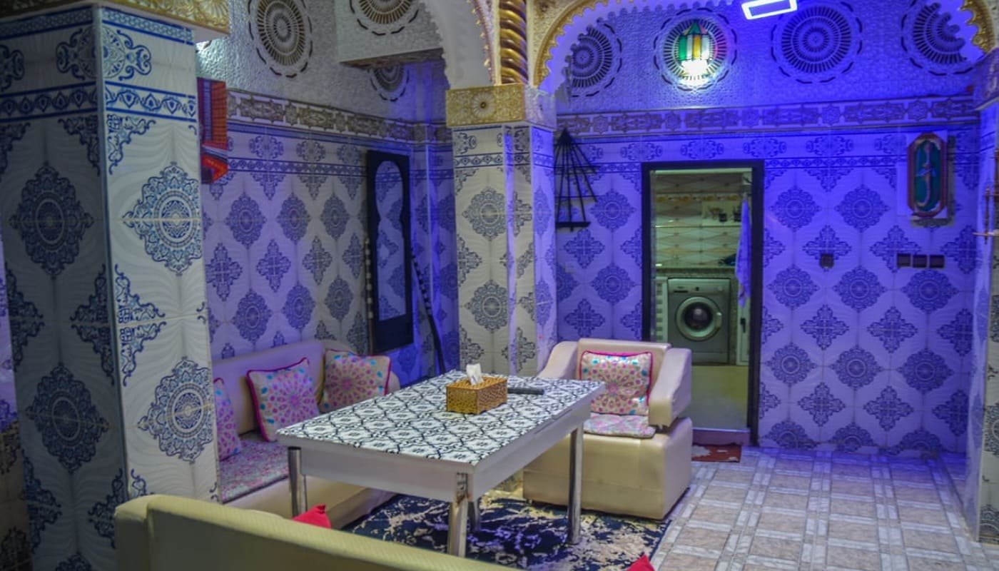 Interior view of LA CASA DAR RAYAN guesthouse showing the elegant staircase with blue and gold decorative railing, ornate pillars, and traditional Moroccan wall carvings in the entryway.