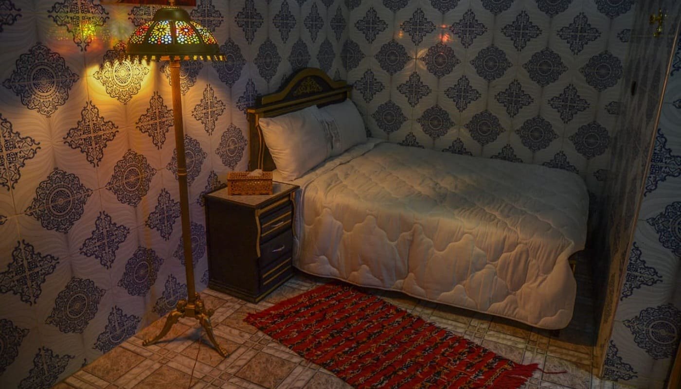 A comfortable bedroom in LA CASA DAR RAYAN guesthouse, featuring traditional Moroccan zellij tiles on the walls, a soft bed, a bedside table with a lamp, and a red rug, offering a relaxing stay in Fes.