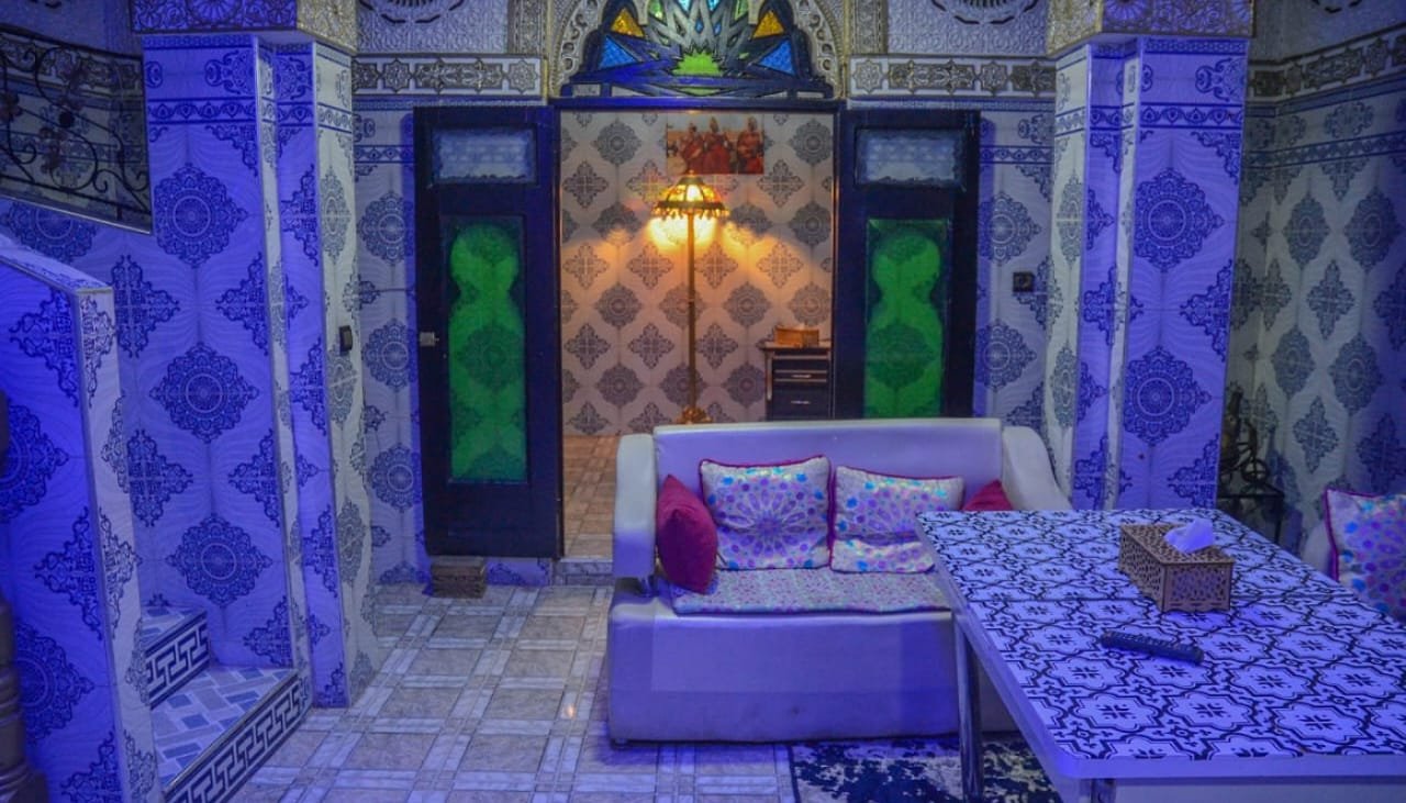 Interior view of LA CASA DAR RAYAN guesthouse showing a comfortable white sofa with pink cushions, a bedroom area with a comfortable bed, and walls decorated with traditional blue and white Moroccan zellige tiles.