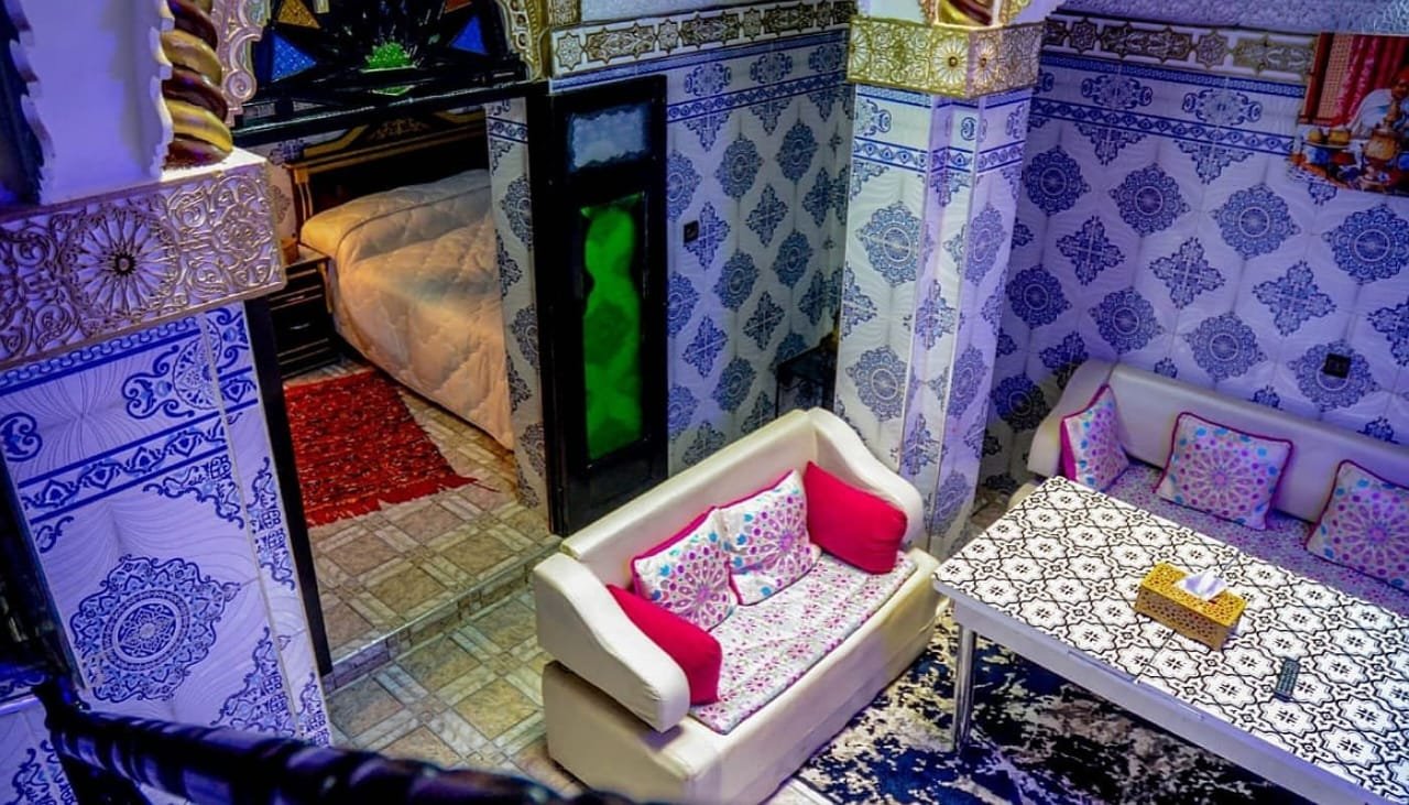 Interior view of LA CASA DAR RAYAN guesthouse showing a comfortable white sofa with pink pillows, a patterned coffee table, and an adjacent bedroom, all surrounded by vibrant blue and white Moroccan zellige wall tiles.