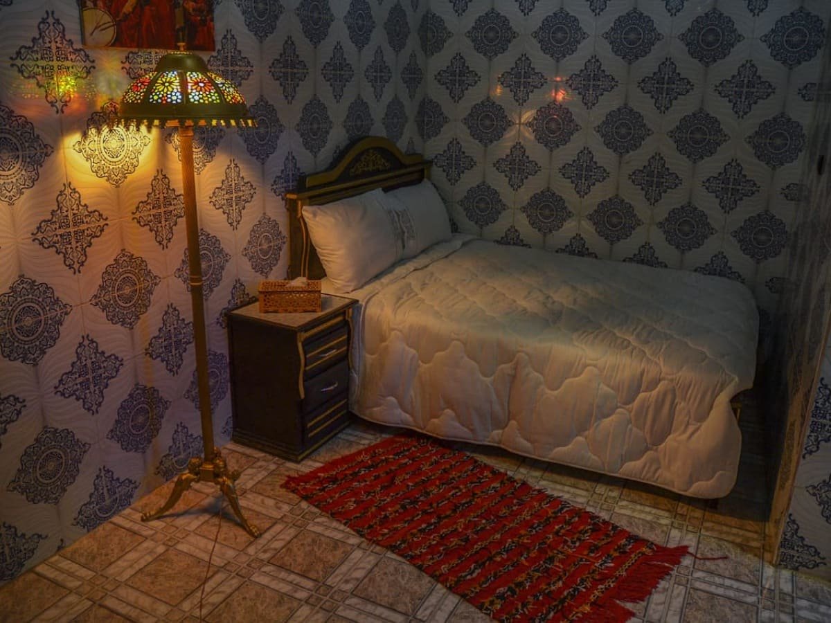 Cozy bedroom in LA CASA DAR RAYAN guesthouse, featuring white bedding, a red rug, an ornate brass Moroccan lantern, and traditional blue and white zellige patterned walls. Ideal Fes Riad room for comfort.