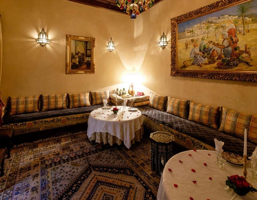 A luxurious traditional Moroccan Riad dining room in Fes, featuring elegant sofas with cushions, ornate wall lanterns, and a beautifully set round table with white linen and rose petals. A large classical orientalist painting hangs on the warm-toned wall, creating a cozy and authentic Ramadan Iftar atmosphere.