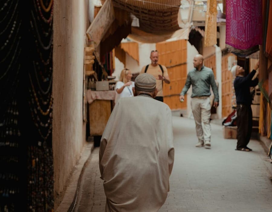 A Moroccan tour guide walking with tourists through a sunlit, narrow alley of Fes Medina during Ramadan. An elderly man in a light-colored Djellaba is seen from behind in the foreground, while colorful textiles and traditional shops line the earthen walls.