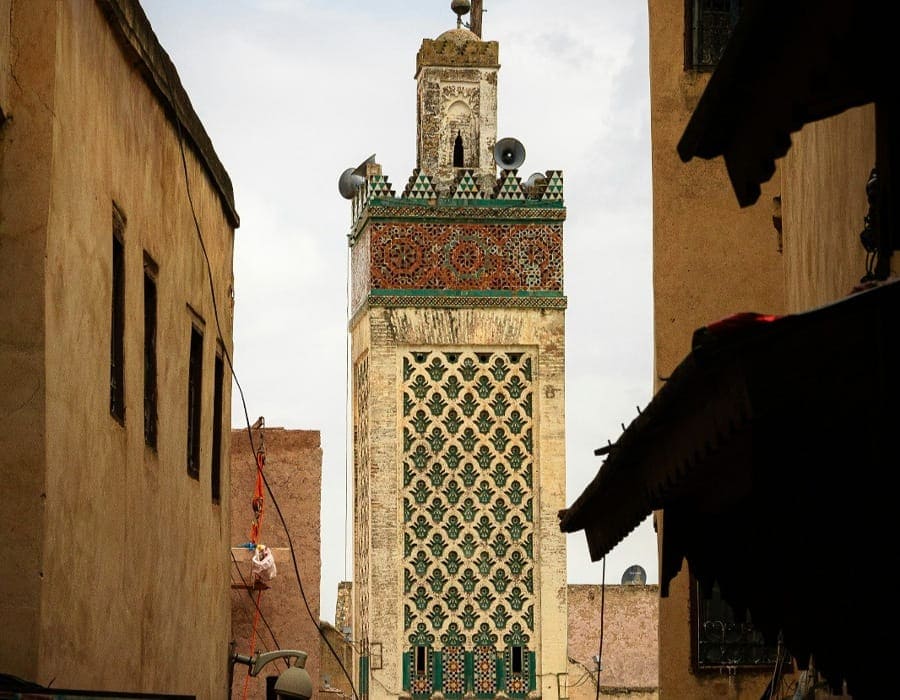 A traditional Moroccan minaret adorned with green and brown mosaics rises between the narrow earthen-walled streets of the Fes Medina during the day. A few old speakers are visible at the top, used for the call to prayer.