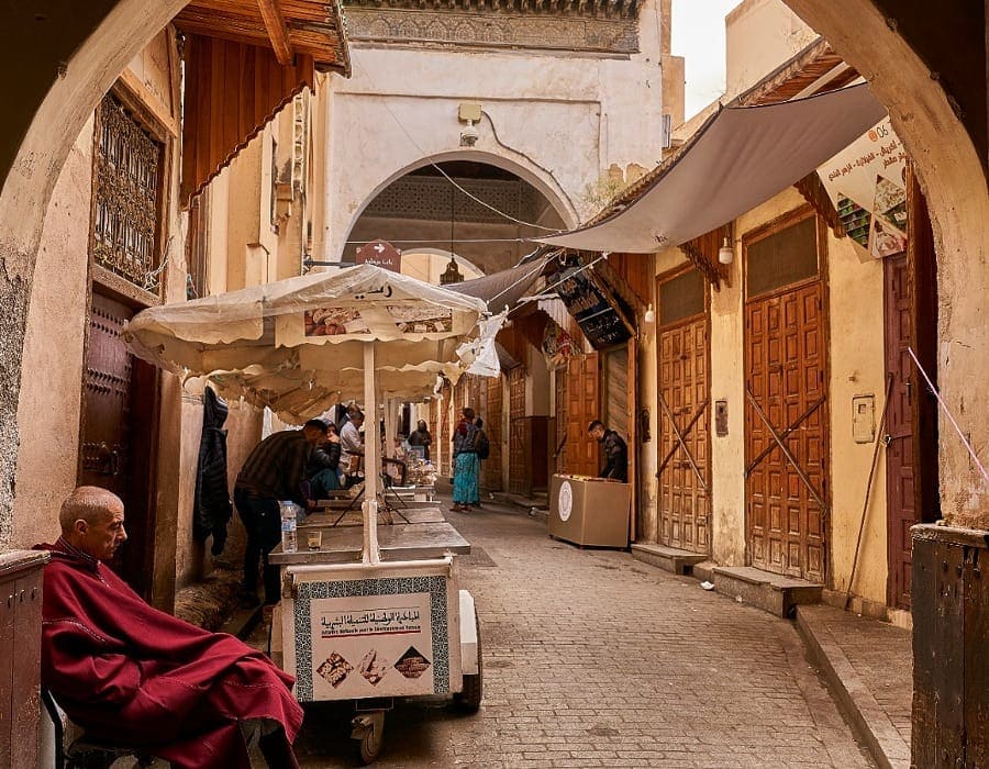 A quiet view of a narrow, cobblestone alleyway in the Fes Medina during the day. An older man in a traditional red Djellaba rests while sitting in front of a small market stall under white umbrellas. The background shows carved arches and traditional wooden doors, perfectly capturing the peaceful atmosphere before the daily Iftar during Ramadan.