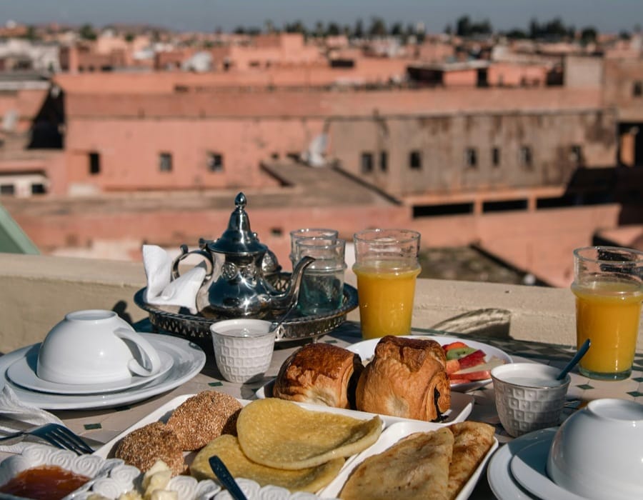 A traditional Moroccan breakfast spread served on a rooftop terrace overlooking the Fes Medina. The table features Moroccan pancakes (Beghrir and Msemmen), fresh orange juice, a silver teapot with glasses, and French pastries, with the reddish-brown earthen city skyline in the background.