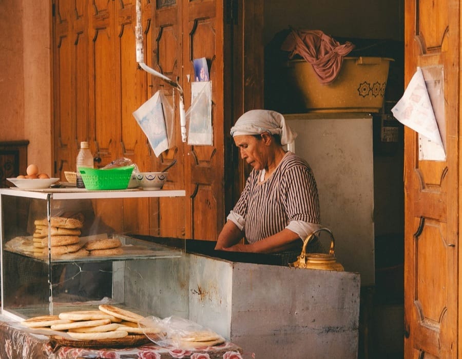 A traditional Moroccan woman artisan wearing a headscarf, preparing handmade flatbreads like Harcha and Msemmen in a rustic Fes Medina bakery. Traditional wooden doors and a vintage brass kettle are visible in the background, capturing the authentic daily life during Ramadan.