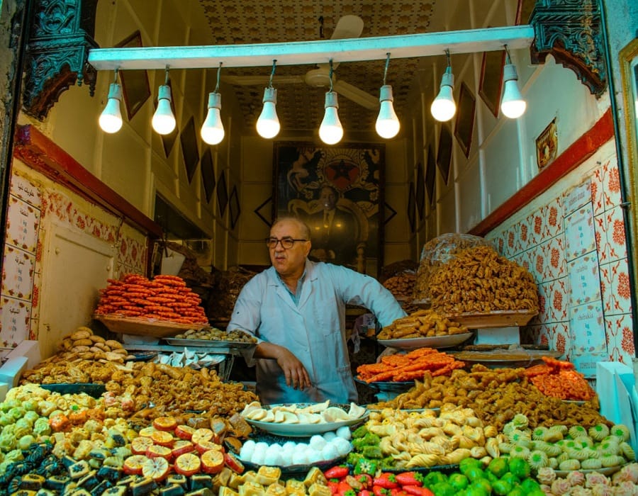 A traditional Moroccan pastry shop in the Fes Medina during Ramadan. A local baker stands behind a counter overflowing with varieties of honey-glazed sweets, including Chebakia, Briouats, and almond pastries. Rows of glowing light bulbs illuminate the vibrant colors of the Moroccan desserts.
