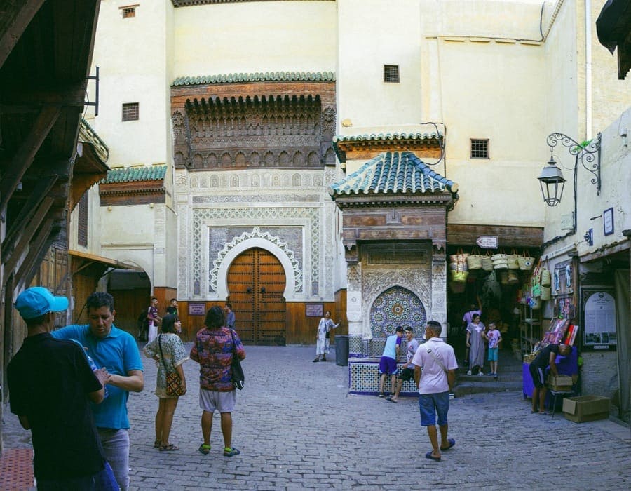 Tourists and locals walking in the Nejjarine Square in Fes Medina. The image features the historic Nejjarine Museum of Wooden Arts and Crafts with its ornate fountain and traditional carved wood architecture during a sunny afternoon.