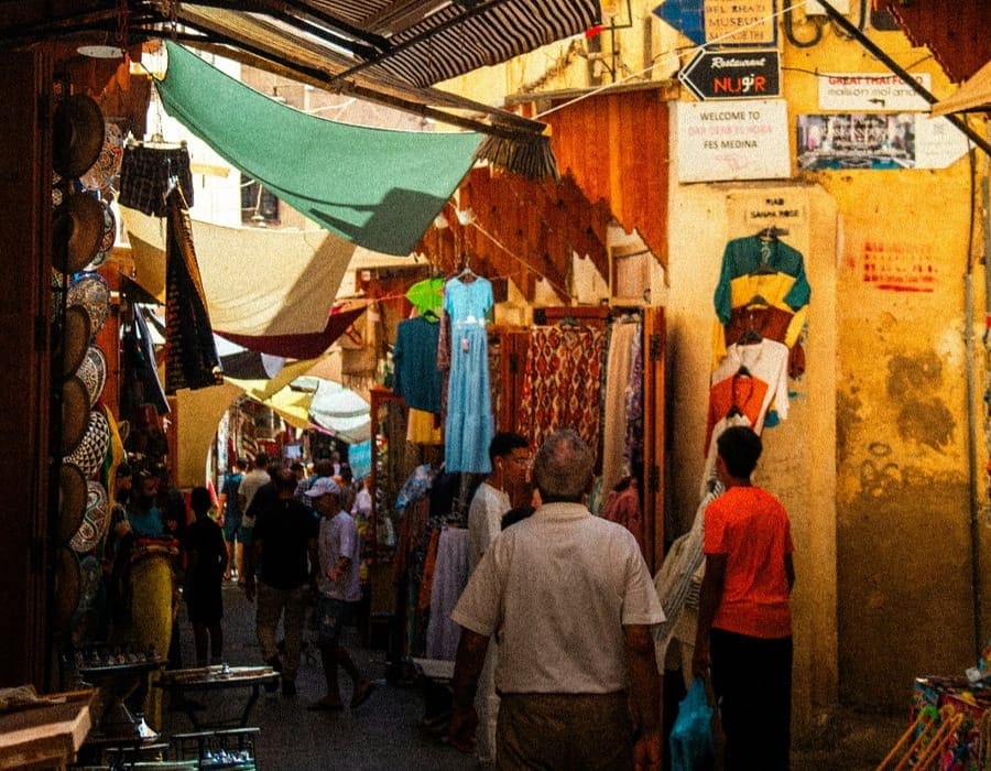 A busy shopping alley in Fes Medina with colorful Moroccan dresses (Kaftyans) displayed on the walls. Tourists and locals walk under cloth canopies, with street signs pointing to local Riads and restaurants.