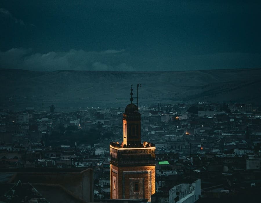 A beautifully illuminated mosque minaret at night overlooking the ancient Fes Medina. The city lights are dim in the background under a dark blue twilight sky, creating a spiritual and serene atmosphere during Ramadan.